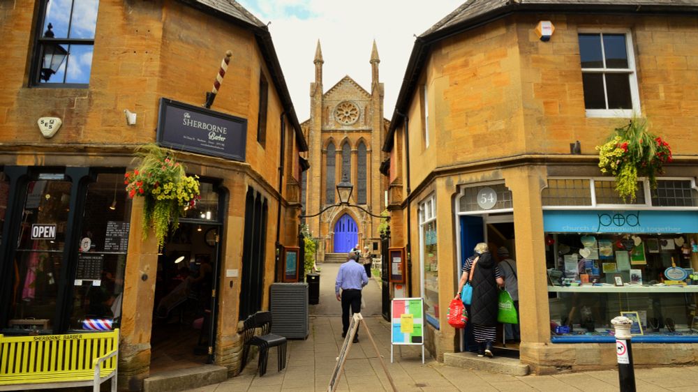 This photo by Jack Greenwood shows a gap between shops on the High Street in Sherborne with a church in the background.
On the left is the Sherborne barber shop with a yellow plastic barrier outside.
On the night is some kind of church community shop.
People mill about. 