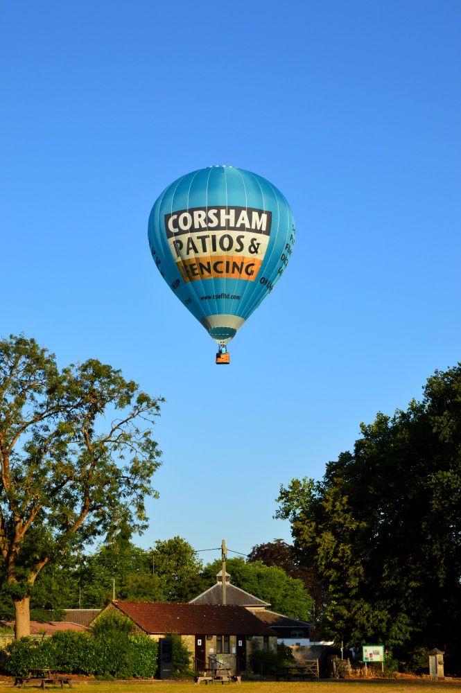 This photo by Jack Greenwood shows a blue hot air balloon above the park in Shepton Mallet.
The sky is completely cloudless.
Trees and small buildings are below. 