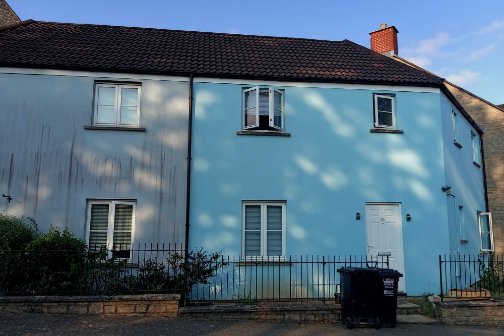 This photo by Jack Greenwood shows a blue terraced old house next to a grey one.
A top window is open and blue sky is above. 