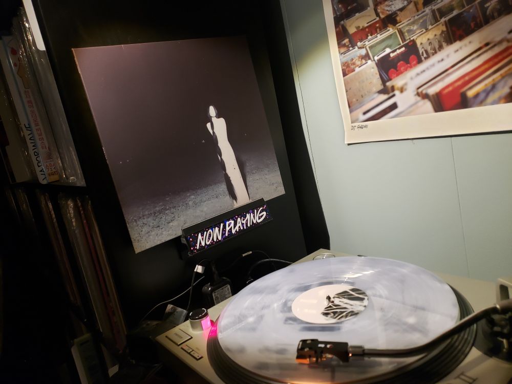 The aforementioned record is playing on a turntable (a milky clear vinyl with white swirls) with the album cover displayed in a "Now Playing" cover holder affixed to a black Kallax shelf full of records 