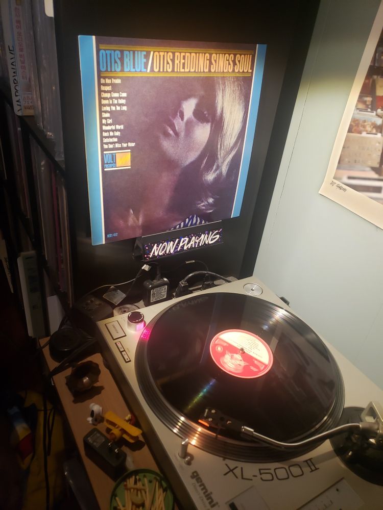 The aforementioned record is playing on a turntable (classic black vinyl) with the cover displayed in a "Now Playing" holder affixed to a black Kallax shelf full of records 