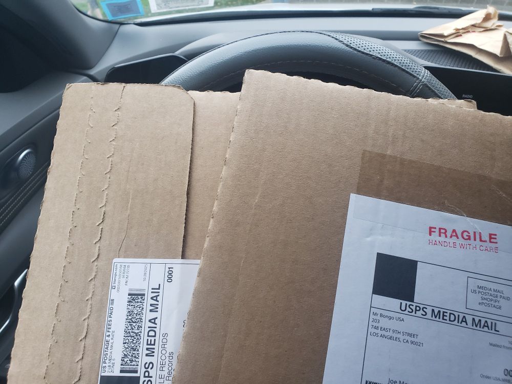 Close up shot of two packages, in the telltale shape of having vinyl in them, over a steering wheel. The vinyls are from Gar Hole Records and Mr. Bongo Records, to give a clue as to what they are!