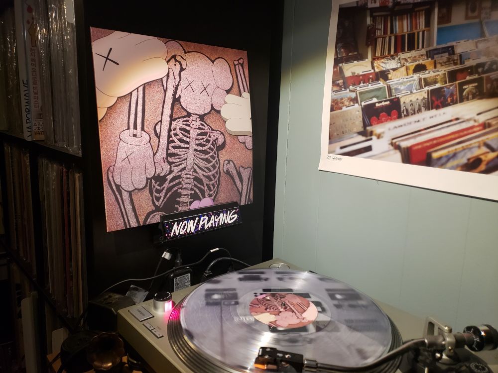 The aforementioned record is playing on a turntable (clear vinyl), with the cover displayed on a "Now Playing" holder affixed to a black Kallax shelf full of records 