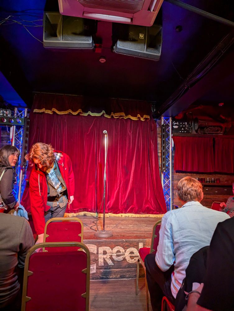 A small performance venue with a red curtained wooden stage, microphone stand. Audience members sit in red chairs awaiting the show.