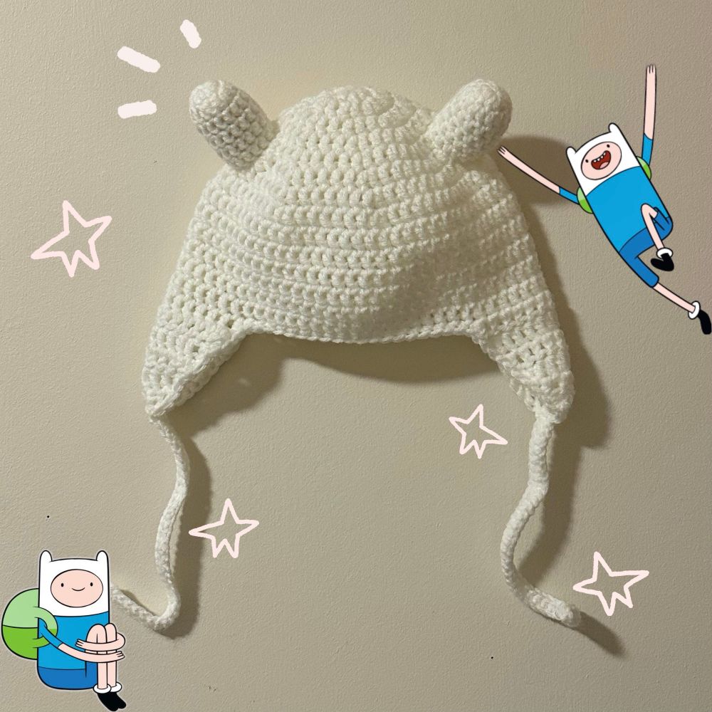Finn the human from adventure time beanie. It is white with Finn’s “ears” at the top of the head, and flaps for the wearers head with strings for tying. Has 4 drawn on stars and 2 pictures of Finn the human in the bottom left and top right corners