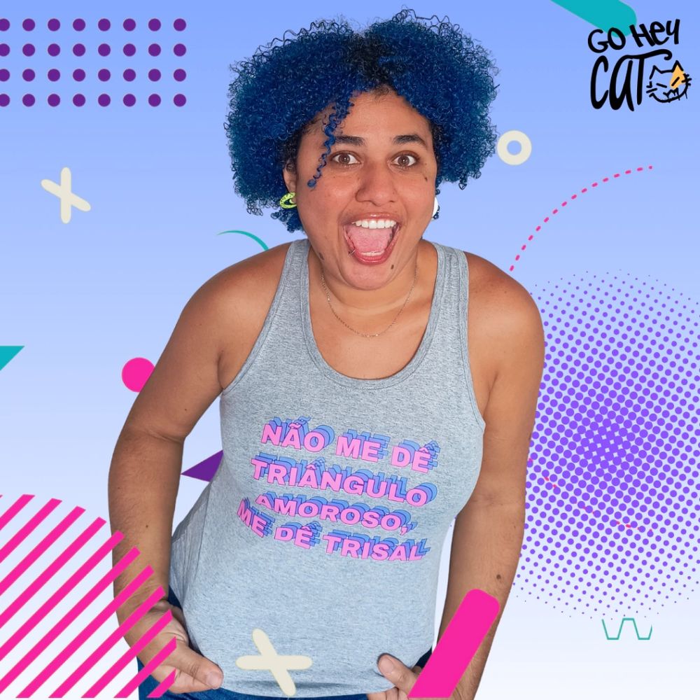 Foto minha sorrindo de boca aberta exibindo a camiseta cinza que estou usando de estampa nas cores da bandeira bissexual (rosa, roxo e azul) escrito em letras maiúsculas: NÃO ME DÊ TRIÂNGULO AMOROSO, ME DÊ TRISAL. Sou uma mulher negra de pele clara, tenho cabelo azul crespo, olhos castanhos e sobrancelhas escuras. O fundo da imagem é um degradê de azul claro com alguns elementos pontilhados em roxo e rosa. No canto superior direito da imagem está o logo da Go Hey Cat, nome da loja por onde é possível comprar essa camisa.