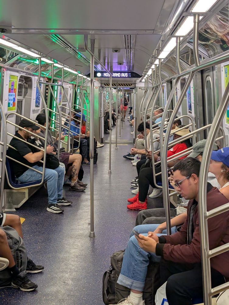 A New York City Subway car with people seated along the sides. Instead of a door at the end of the car, there's an open space, allowing people to move freely and more easily balance the load