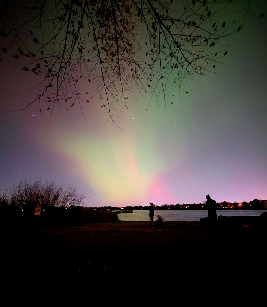 A couple more people on the small Lake Nokomis beach with green, red, and purple hues from the Aurora Borealis mixing with light pollution in the background