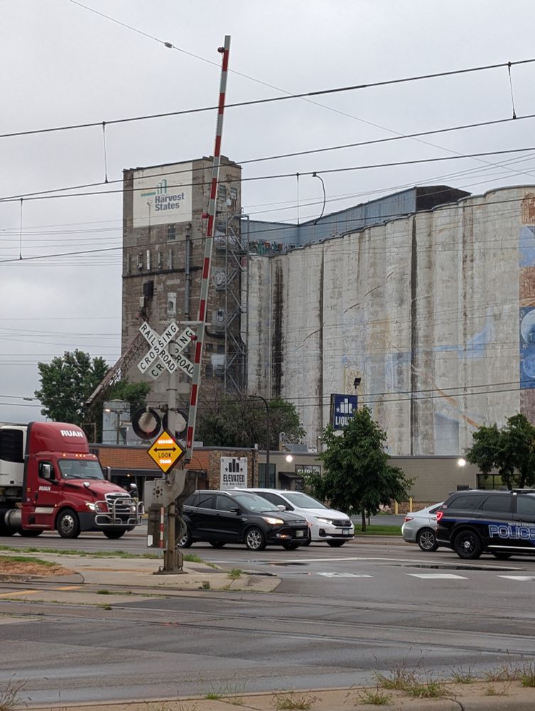 Scene at a grade crossing with a gate arm raised but a yellow diamond "← LOOK →" sign with red "Second train" text lit up. A disused grain silo is in the background and several cars are on the main roadway