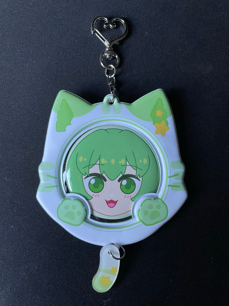 A green and white cat-themed badge protector with a heart shaped key ring at the top and an acrylic charm of a cat tail hanging at the bottom. There is a button pin of Midori’s head in the badge protector