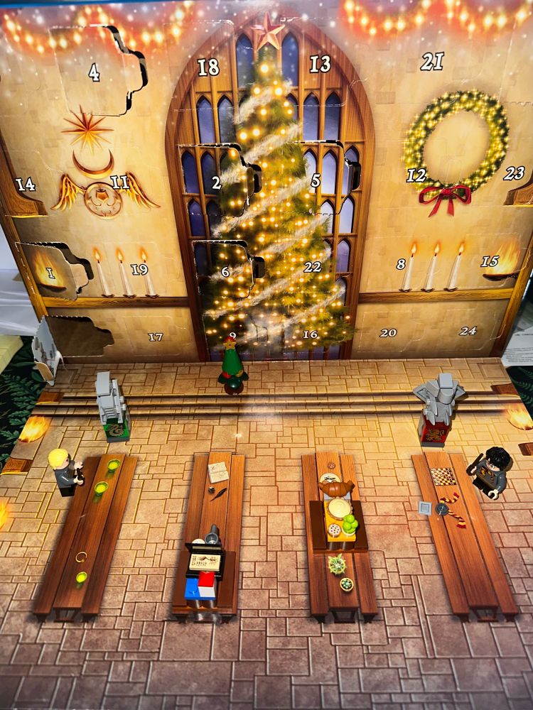 picture of the lego harry potter advent calendar box, set up on its side with the front flap unfolded to show the scene of the great hall of hogwarts. all of the lego builds for each day is shown - harry potter, gryffindor statue, holiday goodies table, table with book, draco malfoy, slytherin statue, and christmas tree
