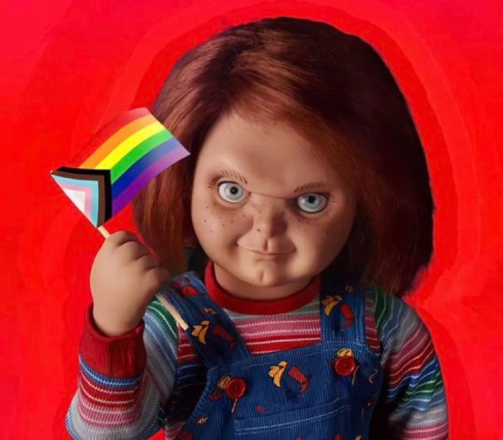 Chucky from the horror movie series stands against a bold red background, smirking mischievously while holding a small Progress Pride flag. The doll wears striped sleeves and blue overalls with colorful patterns.