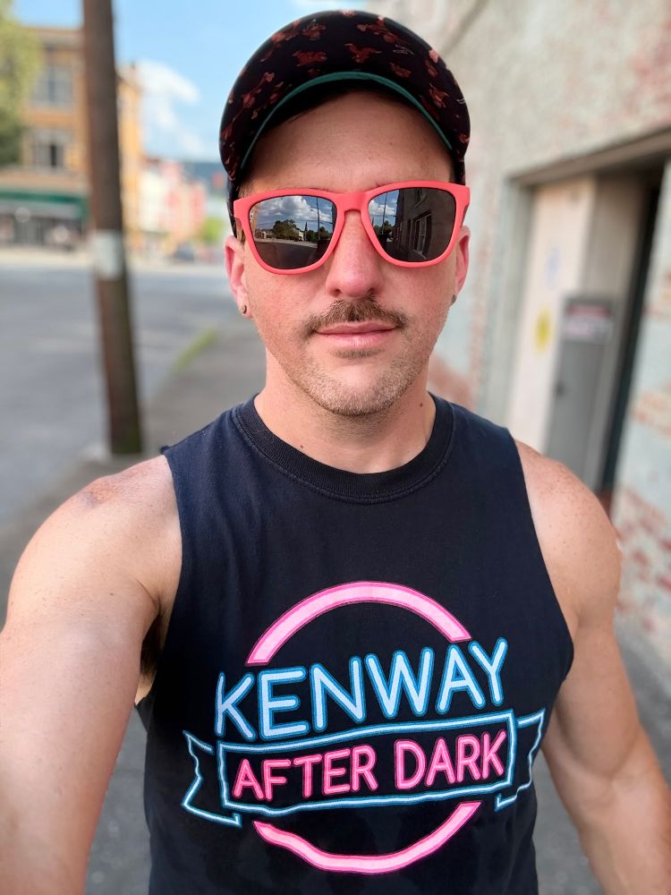 A person wearing a black sleeveless shirt with the neon-style text “Kenway After Dark” in blue and pink lettering, coral sunglasses, and a dark baseball cap stands outdoors on a city street, taking a selfie.