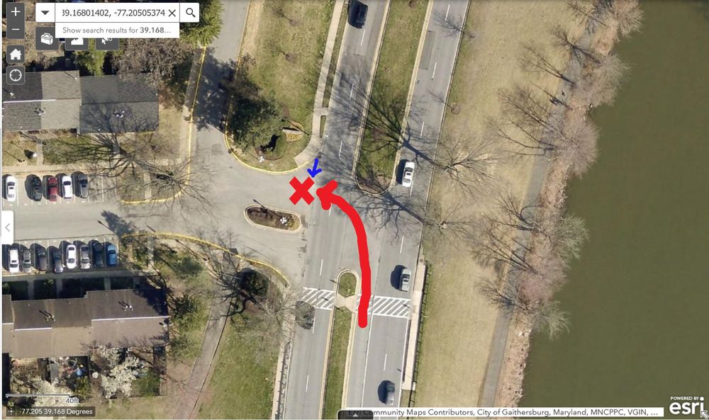 Satellite image of the T intersection of a very wide two-way driveway to a complex of attached houses, and a four-lane divided road with a left turn only lane. The south side of the intersection has a marked crosswalk leading to a bus stop. The crosswalk across the driveway is not marked. There is a red X in the north part of the driveway, on the west side of the intersection, indicating the location of the crash. There is a red arrow pointing north and west from the left turn only lane toward the red X, indicating the path of the driver. There is a blue arrow pointing south toward the red X, indicating the direction of the bicyclist, who might have been riding in the road or might have been riding on the sidewalk.