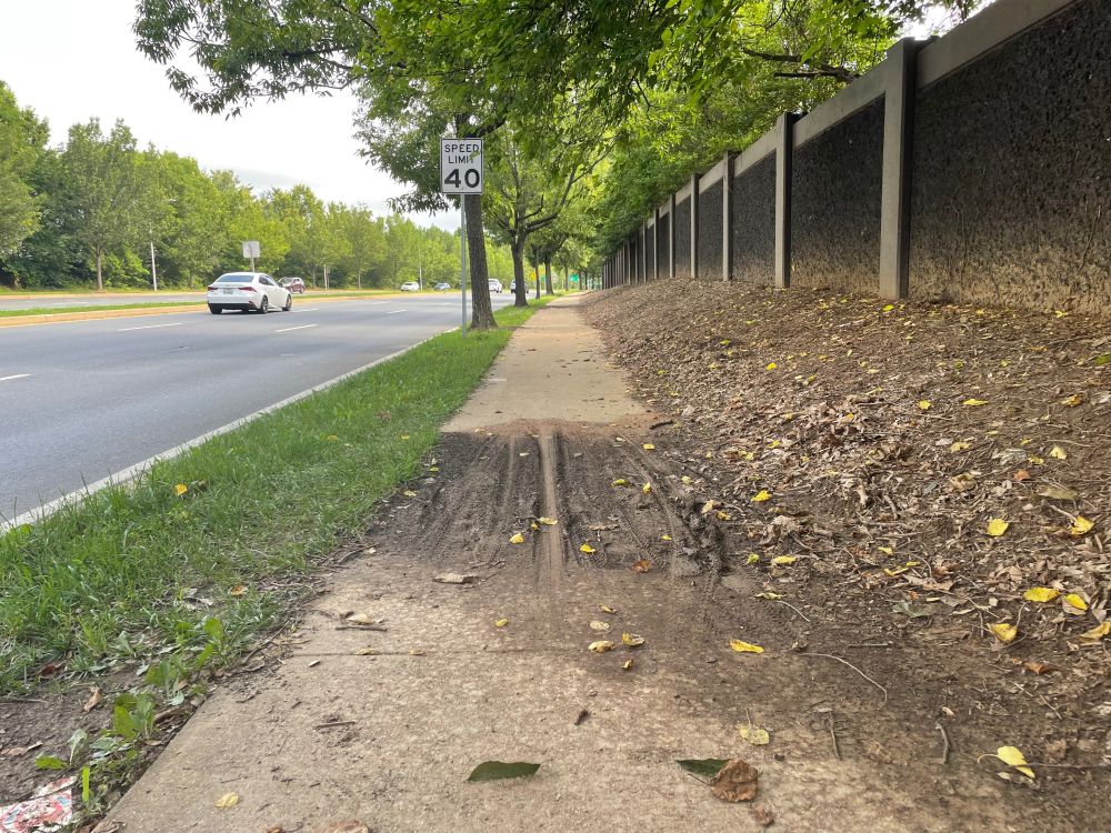 A muddy patch in a sidewalk, with lots of bicycle tire tracks, next to a six-lane divided state highway with a 40 mph speed limit sign. On the other side of the sidewalk is a berm and a low sound wall to protect people in a residential area from the effects of cars that aren't theirs.
