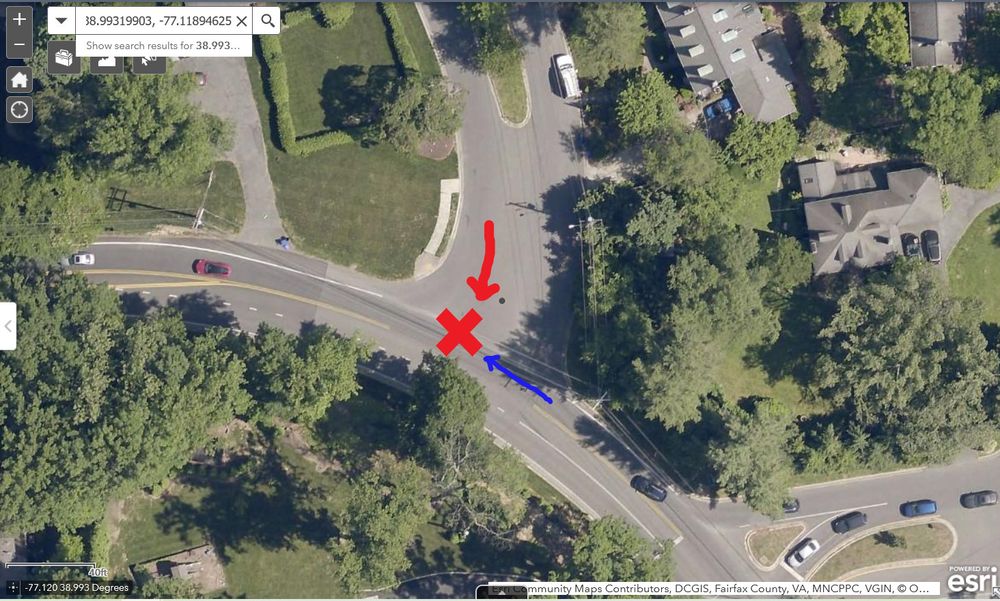 Satellite image of the T intersection of a very wide side street with a narrower two-lane state highway. There is a red X in the westbound lane of the state highway, indicating the location of the crash. There is a red arrow pointing south in the very wide side street toward the red X, indicating the path of the driver, who had a stop sign. There is a blue arrow pointing west toward the red X in the westbound lane of the state highway, indicating the path of the bicyclist.