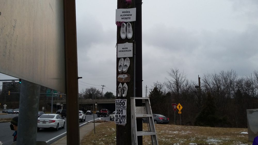 Three ghost shoes and plaques, on a utility pole, next to a bus stop, next to a highway interchange.