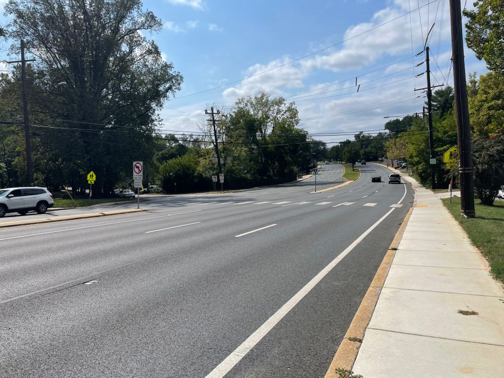 An uncontrolled marked crosswalk across seven lanes of high-speed state highway, looking downhill. There are unbuffered sidewalks on both sides. There are school crossing signs at the marked crosswalk.