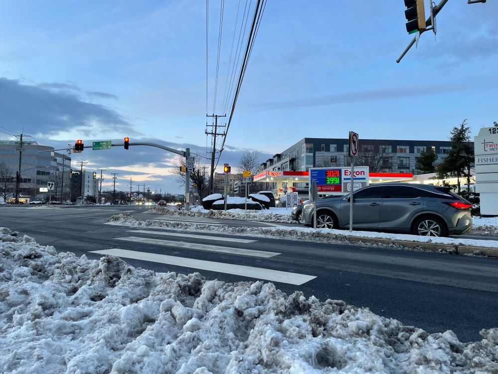 A large signalized intersection of a stroad, where the snow has been cleared from the road down to bare asphalt, and the places where people are supposed to walk are piles of slush and ice that people have had to walk through.