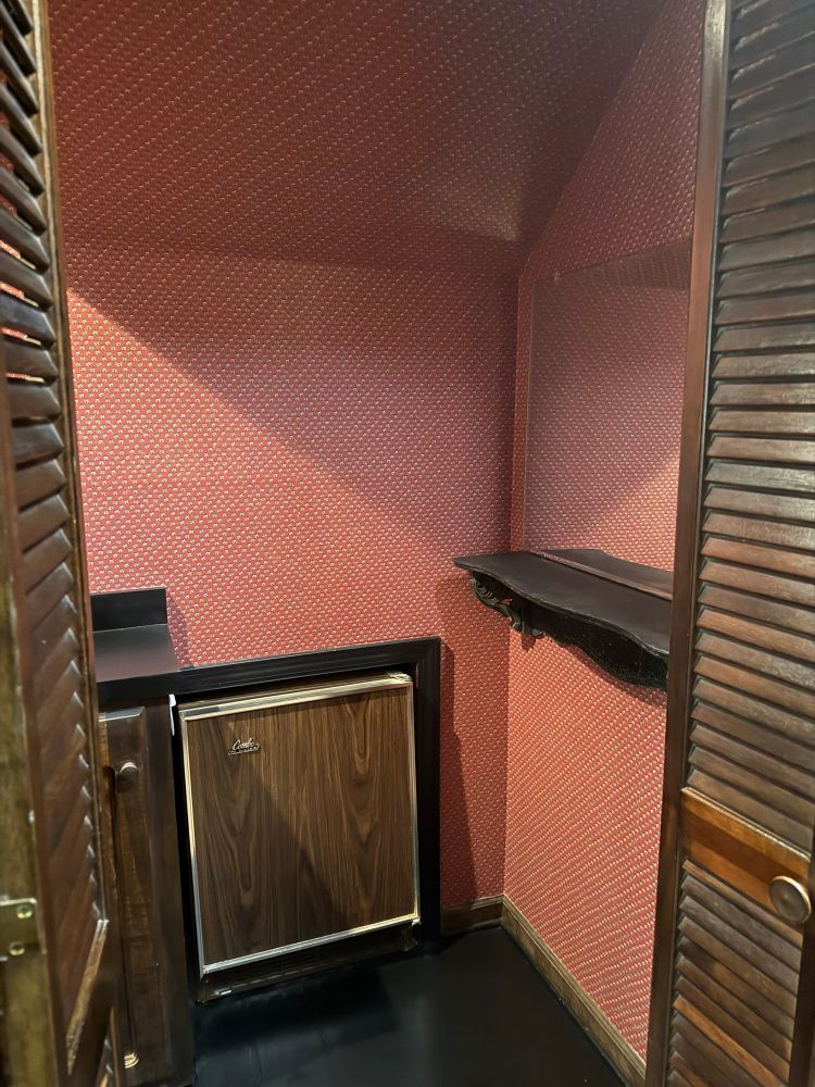 Before image showing the right half of the room with a shelf under the mirror, dark wood accordion doors, and a small old-fashioned beverage refrigerator directly under the sloped roof.