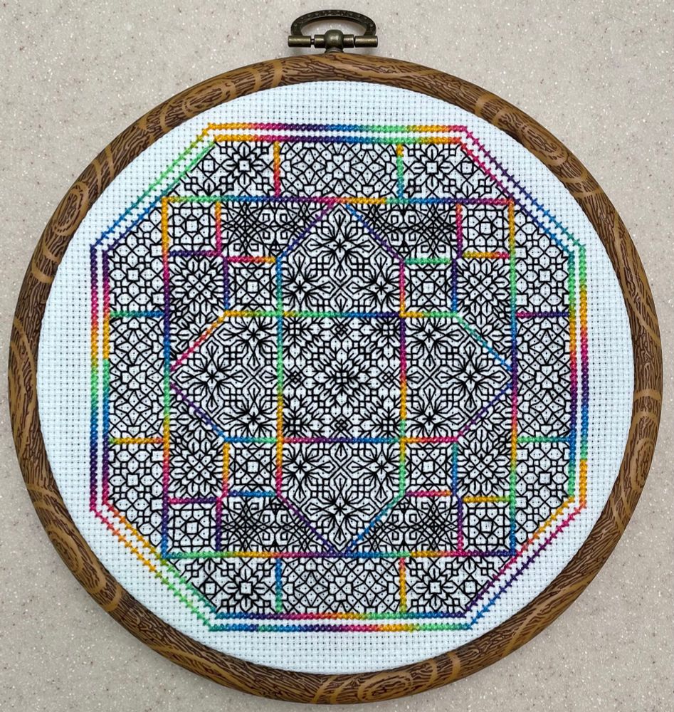 Intricate blackwork that looks a little like the outlines for a stained glass window, coupled with rainbow cross stitch that borders the design. Framed in a wood-looking circular embroidery hoop
