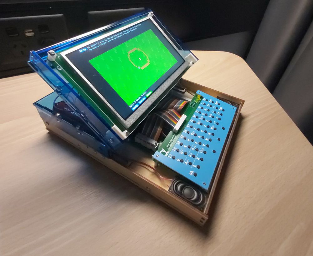 An assembled Agape computer kit sitting on one of those desks that's supposed to look like wood grain but obviously isn't. The base of the computer itself is a box made of wood though! The top half of the case is transparent blue acrylic and the keyboard is also blue. Little speakers are mounted on either side of the keyboard. The computer is switched on and the screen displays a cheerful grassy world with a pixel art-adjacent aesthetic. There are some wires and lovely rainbow ribbon cables visible here and there, the overall effect being of a delightfully hardware hacker-ish netbook.