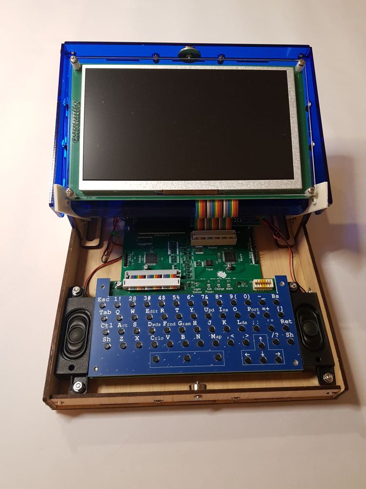 An Agape computer kit: the case is wooden and contains visible circuit boards and a keyboard with a blue overlay to label the buttons with speakers on either side of it. The lid is translucent blue acrylic and contains a screen that connects to one of the circuit boards with a pretty ribbon cable. Every month is Pride month here.