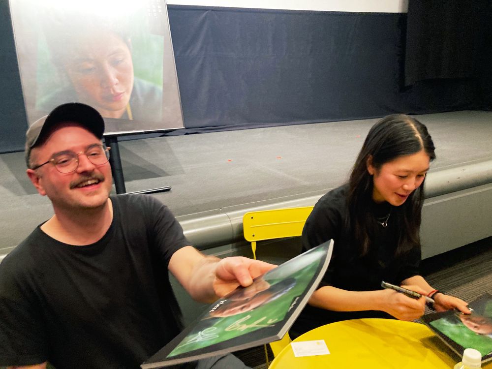サイン入りの映画パンフレットを手渡すバス・ドゥヴォス監督、サインを書いているリヨ・ゴンさん