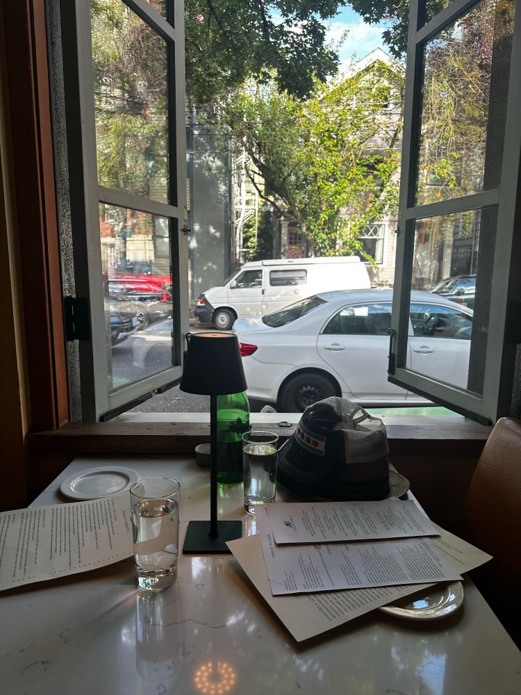Table for two at Serrato’s in Portland. The table top is marble. An open window overlooks NW 21st Avenue. Hats are on table as are menus.