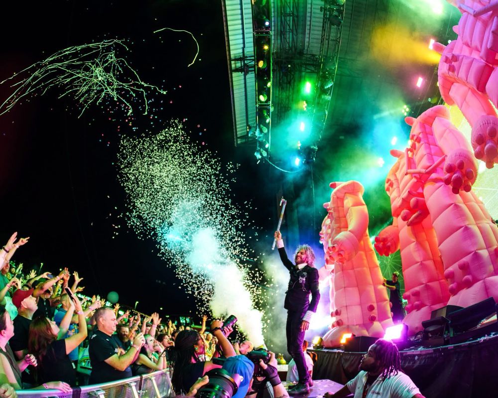 Wayne Coyne of the band The Flaming Lips stands on an amp. There are giant pink blow up robots behind him on stage, the audience in front, and confetti being fired from cannons 