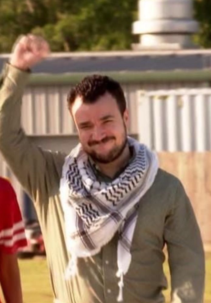 Photo of a young smiling man with dark mustache and beard, dressed in a green long sleeved shirt and keffiyeh, raising his right fist in the air  