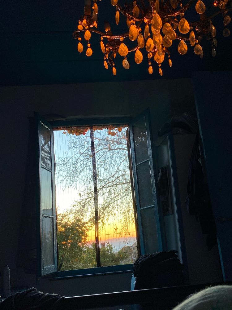 Greek cottage, view from bed, looking through blue shuttered window at sunrise over the sea in the distance! Old chandelier not switched on, just reflecting orange glow! 😍