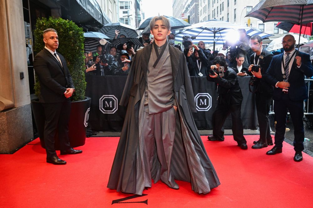 full body photo of scoups outside the met gala in a multilayered grey hanbok-inspired suit including a floor length outer layer. he has his hands in his pockets and he lools confident and almost cocky. it is sexy