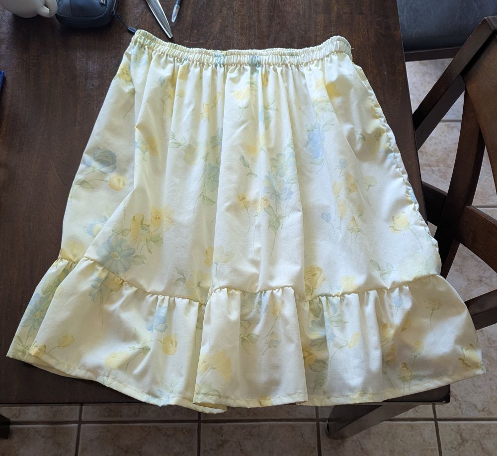 A two-tiered skirt made of pale yellow fabric that has a sort of vintage print of blue and yellow flowers. It's laid out flat on a kitchen table.