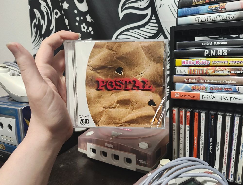 A sealed copy of Postal being held up in front of my Dreamcast and some games. 