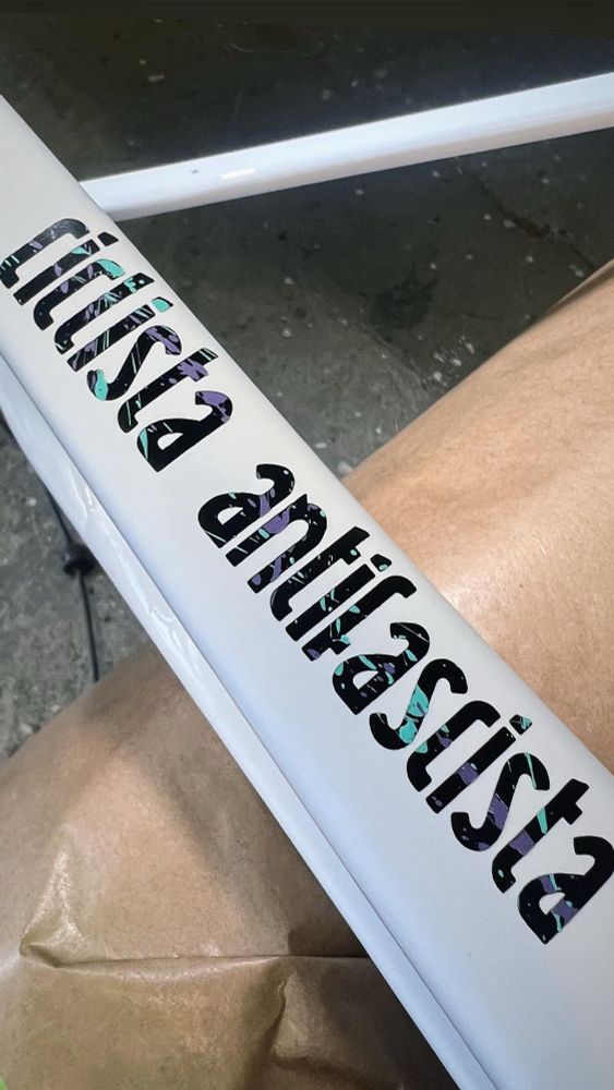 Bild eines Rahmens mit dem Schriftzug ciclista antifascista der Rahmen ist weiß der Schriftzug ist schwarz mit lila und celesten sprenkeln 