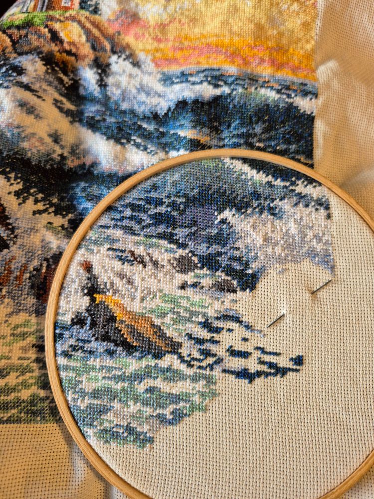 A counted cross stitch project in progress, featuring a lighthouse on a cliff over the ocean at sunset.