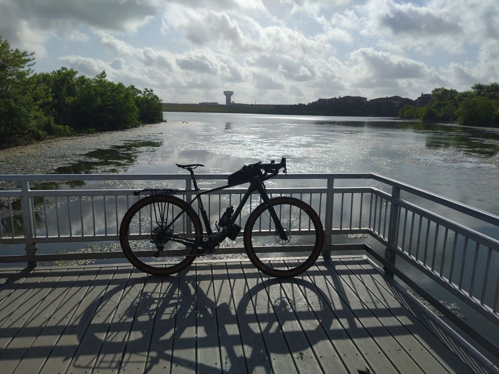 A bicycle leaning against the rail on a short pier, casting a shadow from the mid-morning sun. A lake with trees is in the background. The sun is brightly shining through puffy, white clouds.