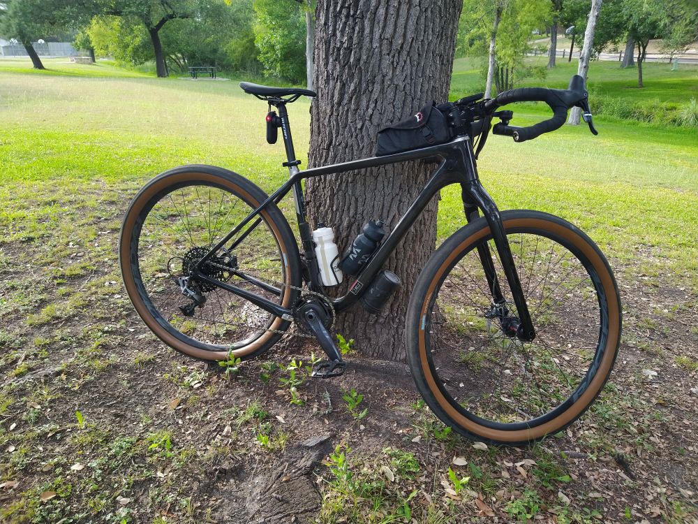 A black Salsa Cutthroat bicycle leaning against a tree, with a park and stream in the background.