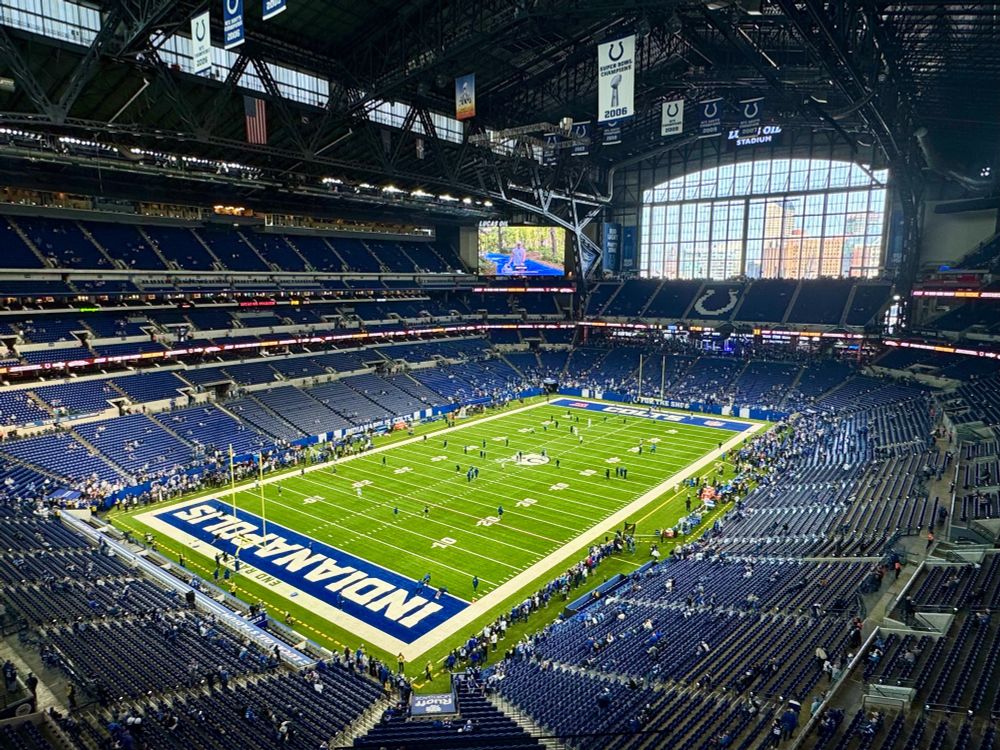The Indianapolis Colts stadium. A large window is in the background, with a green football field and thousands of blue seats surrounding the field. 