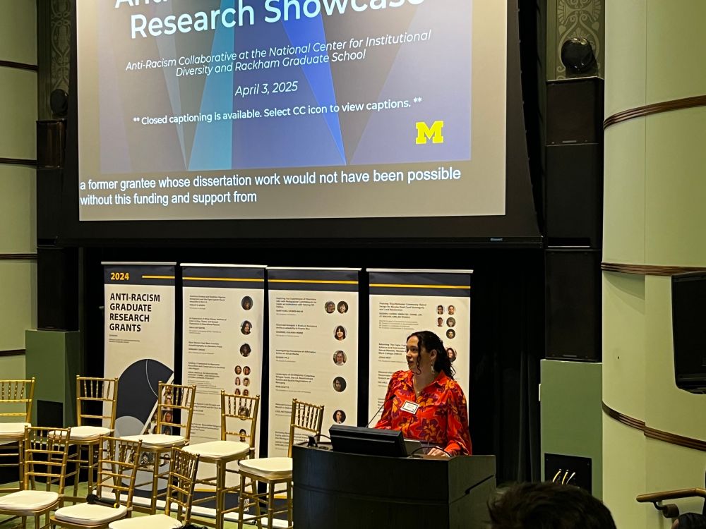A photo of me speaking behind a podium. I am wearing an orange floral blouse and there are gold chairs to my left for the grantee Q&A panel. 
