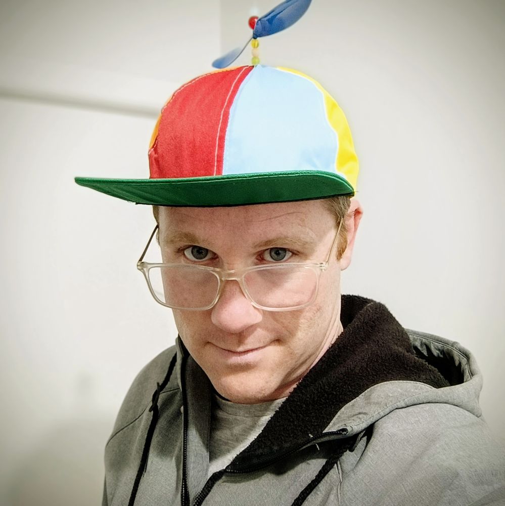 A pale male is appears not stale by virtue of the red, green, yellow and blue hat they wear with a propeller on top. They are also wearing a boring gray jacket, and clear framed glasses. They look into the camera over the frame of the glasses.