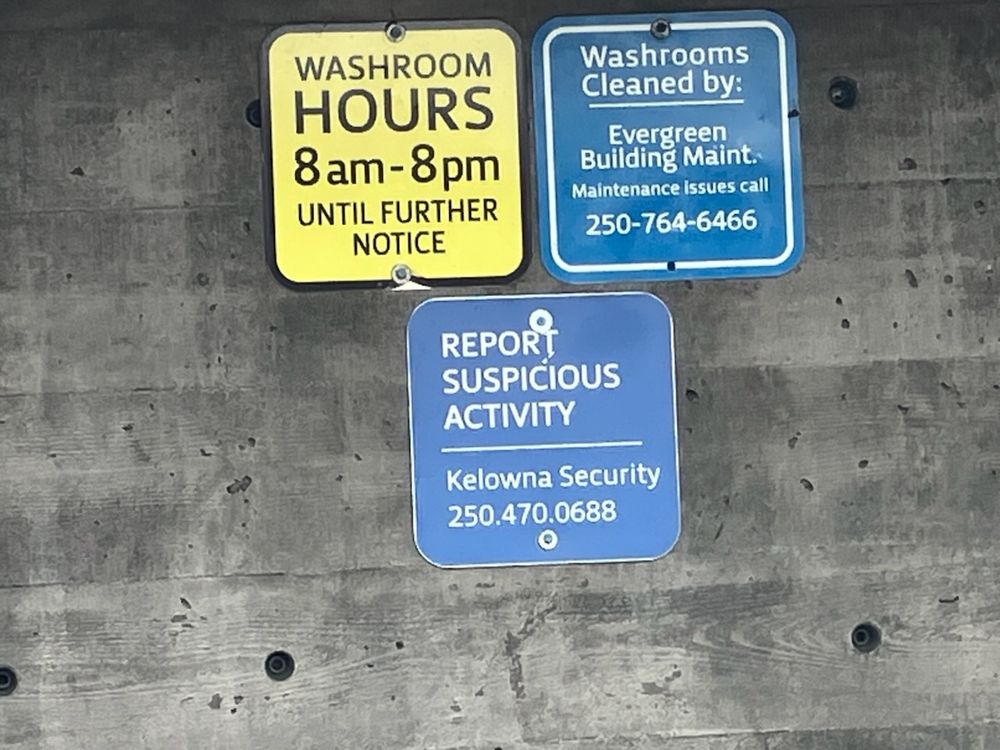 This is a picture of three signs on a grey wall. The first sign is yellow with black lettering and has the text "Washroom Hours 8am-8pm until further notice". The second sign is blue with white lettering and reads "Washrooms cleaned by Evergreen Building Maint. Maintenance issues call 250-764-6466". The third sign is blue with white lettering and reads "Report suspicious activity. Kelowna Security 250-470-0688."