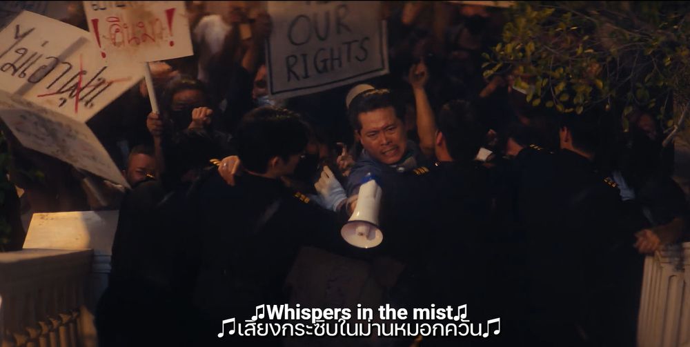 Screenshot of the thai serie "The Next Prince", episode 10.
We see a demonstration of the people, with placards, one of which speaks of civil rights, held back by guards. A man emerges of it, with a megaphone and an expression of anger and determination on his face.