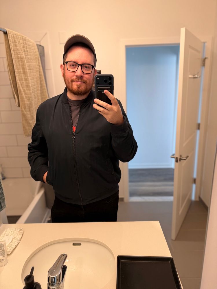 A man stands in a well-lit bathroom, taking a selfie with a smartphone. He has a beard and is wearing glasses, a black cap, and a black bomber jacket over a dark shirt. His hands are casually placed on his hips. The bathroom features a white sink with a modern chrome faucet, and a soap dispenser is visible on the counter. To the left, a white tiled bathtub is partially visible, with a beige towel hanging on a rack above it. The walls are painted a light color, and a wooden door with a silver handle leads to another room, which appears to have a light blue wall and a wooden floor. The overall atmosphere is clean and contemporary.