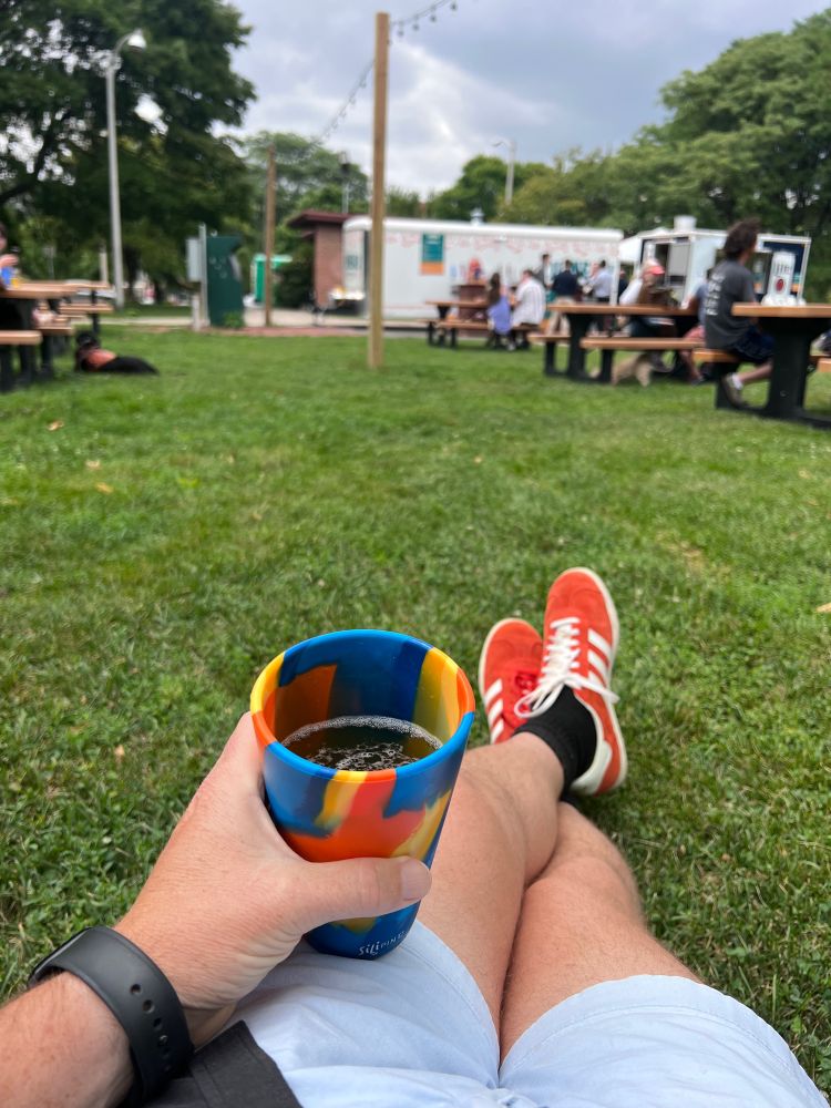 This is a picture of my point of view. Visible in the foreground is a blue, yellow, and orange cup that has beer in it. I’m holding it. In the background, picnic tables, grass, gray sky, and a white mobile trailer with beer taps on it.