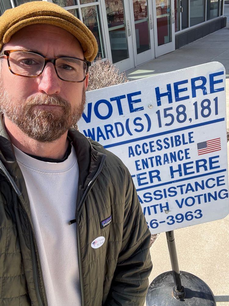 White bespectacled bearded man wearing a green jacket with “I voted” sticker. He stands in front of a “vote here” sign. 