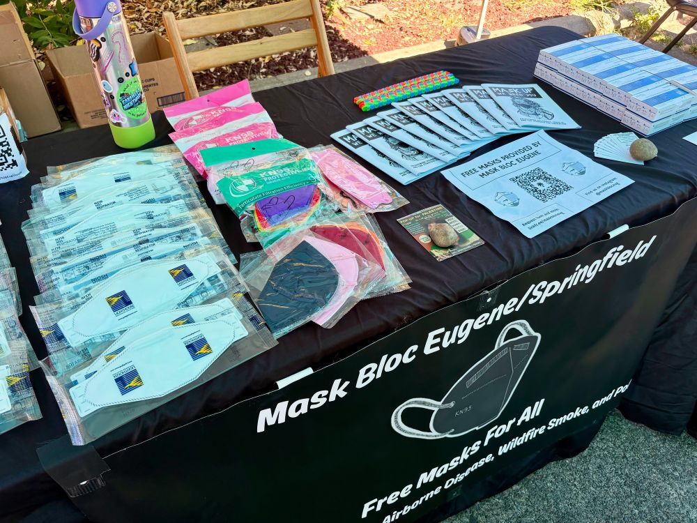 Mask Bloc Eugene’s table outside, with a black tablecloth and a banner in front with a grey KN95 on it. There’s rows of respirator masks, flyers for an upcoming event, a QR code to our links, business cards, zines, fidget toys, and stacks of rapid tests.