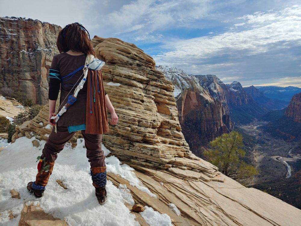 Kathleen Rowan, in a post-apocalyptic costume complete with (toy) archery bow inspired by Horizon Zero Dawn, stands boldly in the snow atop Angel's Landing at Zion National Park in Utah, USA. The canyon is so far below that the river looks like a road and the road is barely visible.
