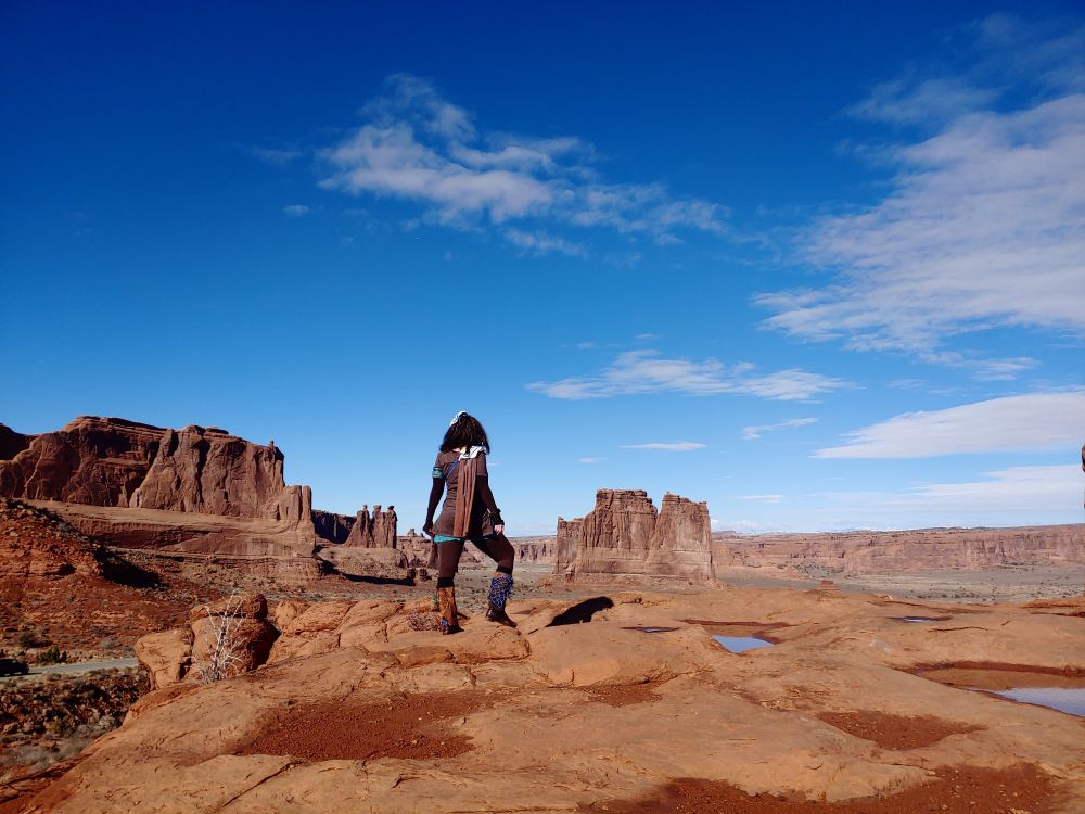 Photo: an adventuress pauses to survey the wide, orange and blue vista of natural desert monoliths under open sky. She is dressed as a Nora from Horizon Zero Dawn, in brown with accents of blue and a curved white "machine plate" clasping a short cape to one shoulder. Puddles of water pooled in shallow hollows in the plateau on which she stands reflect back the azure of the sky.
Arches National Park, Utah, USA.
Photo by my husband.
